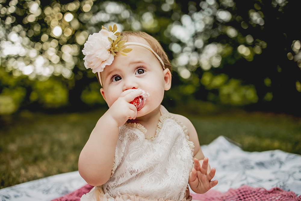 At a first birthday photo session a baby girl eats a strawberry off of her cake and a cake smash picture session